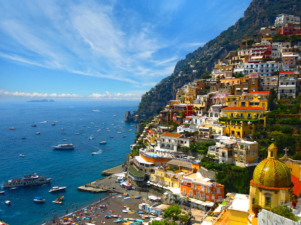 Colorful buildings cascade down a steep hillside to a crowded beach with boats on the water under a bright blue sky.