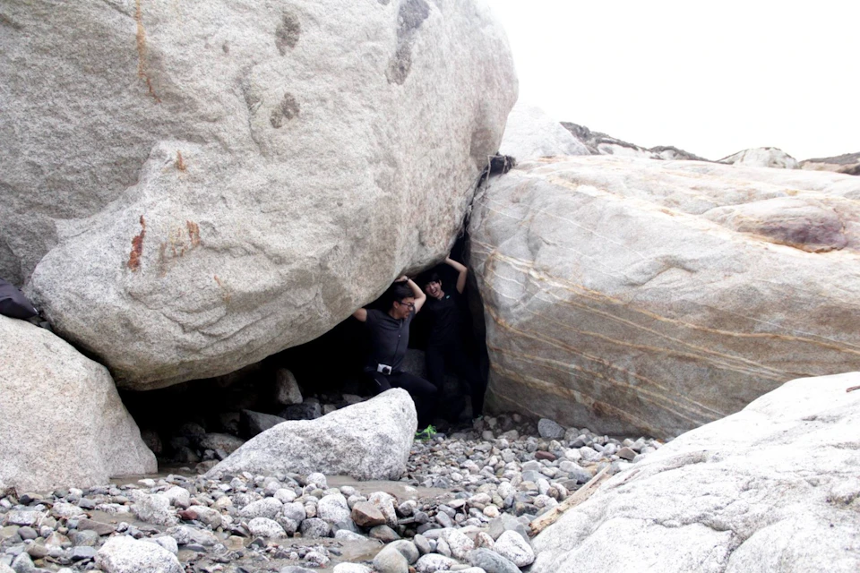 Two individuals appear to be playfully pretending to hold up a large boulder in a rocky outdoor setting.