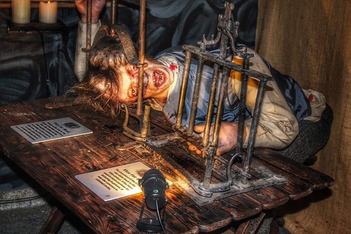 Figurína připomínající mučeného člověka se zoufalým výrazem je uvězněna ve staré kovové mašinérii na dřevěném stole.