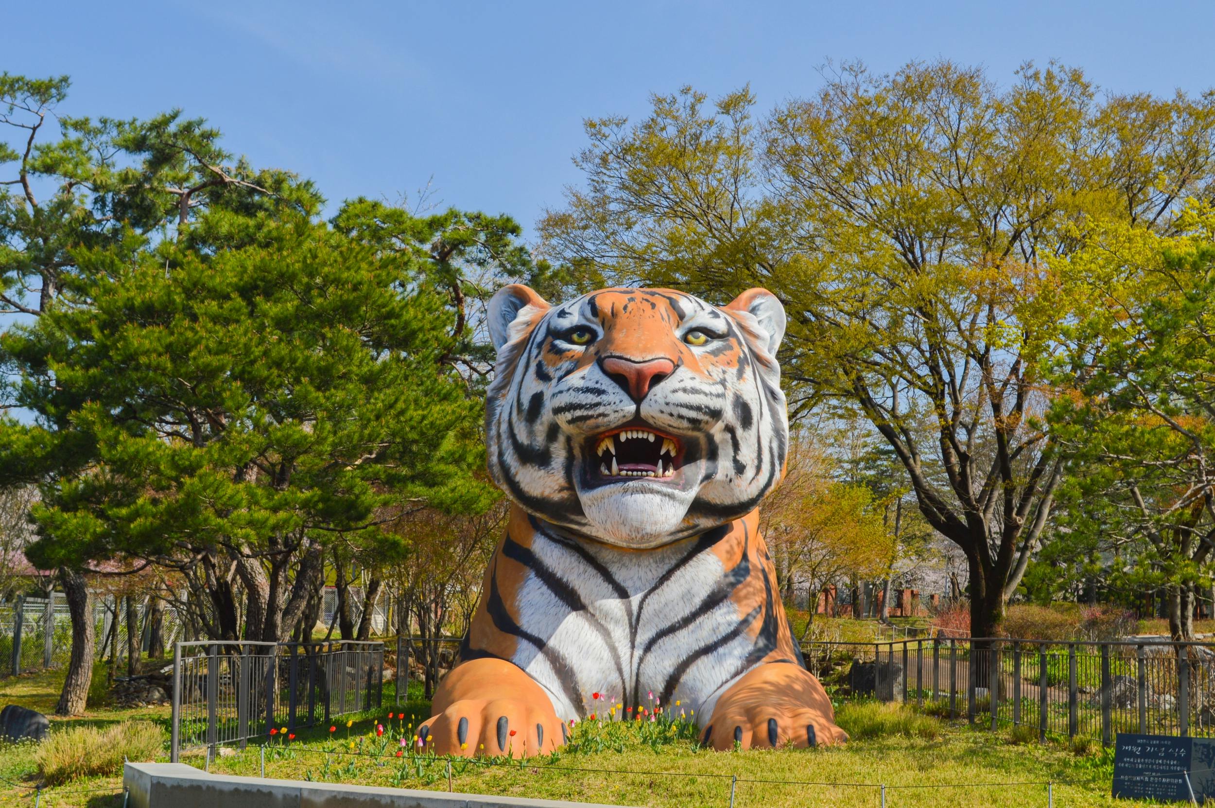 A large tiger sculpture in a park surrounded by green trees and flowers under a clear blue sky.