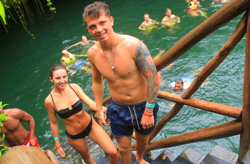 Una dona amb bikini negre i un home tatuat amb banyador blau somriuen mentre pugen escales des d'una piscina natural; altres neden de fons.