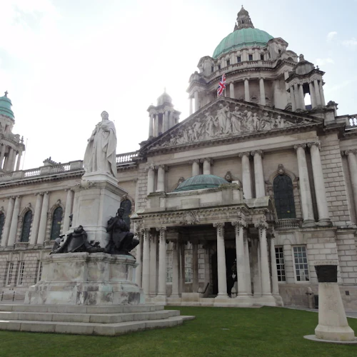 Historic building with ornate columns and domes, a statue of a robed figure with two seated figures in front, and a Union flag.