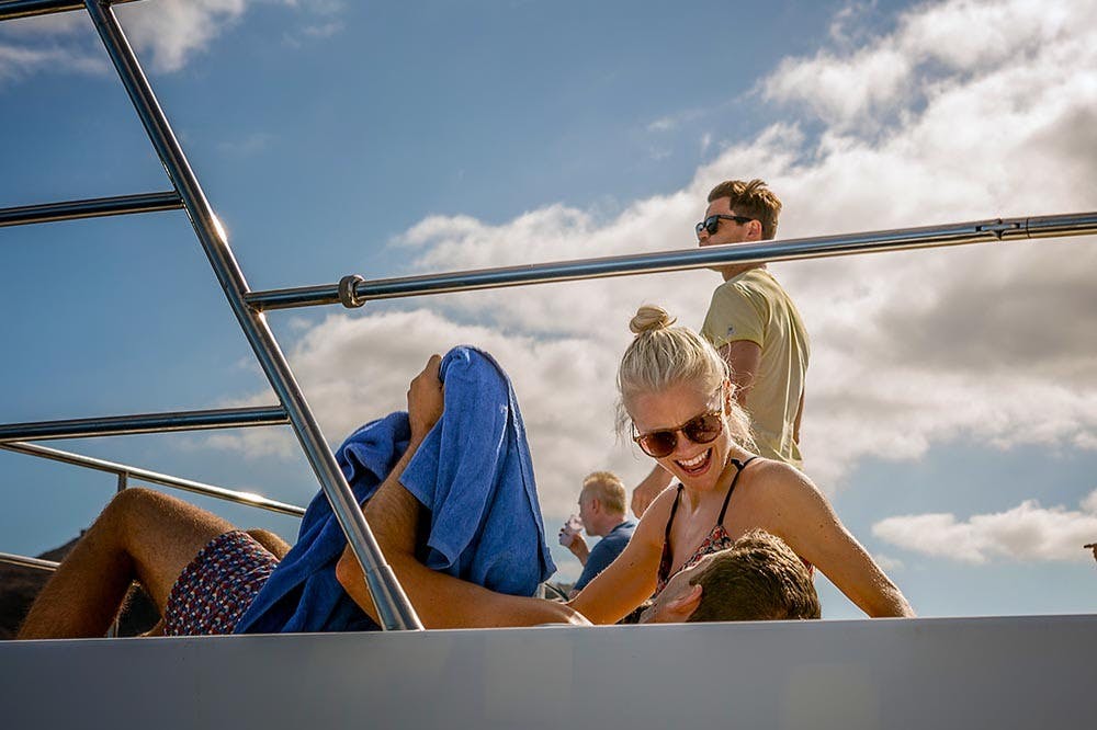 船上有三个人：一个戴太阳镜的女人微笑着，一个戴太阳镜的男人站着，另一个戴着蓝色毛巾的男人躺着。