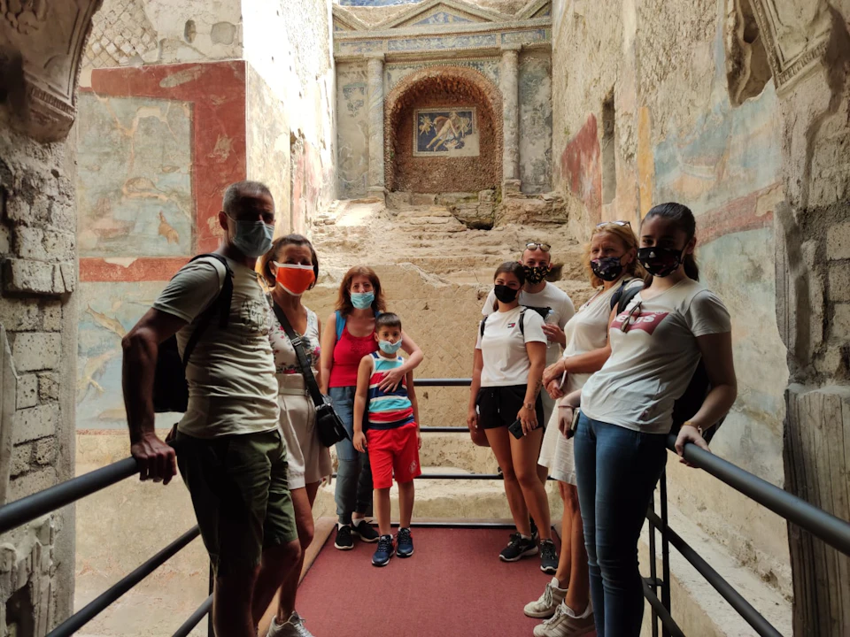 A group of eight people wearing masks standing on a pathway inside an ancient, mural-decorated ruin.