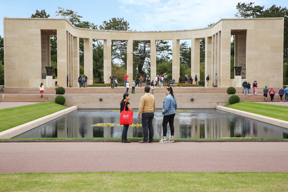 People walking and talking in front of a memorial structure with columns and a pool, surrounded by trees and grass.