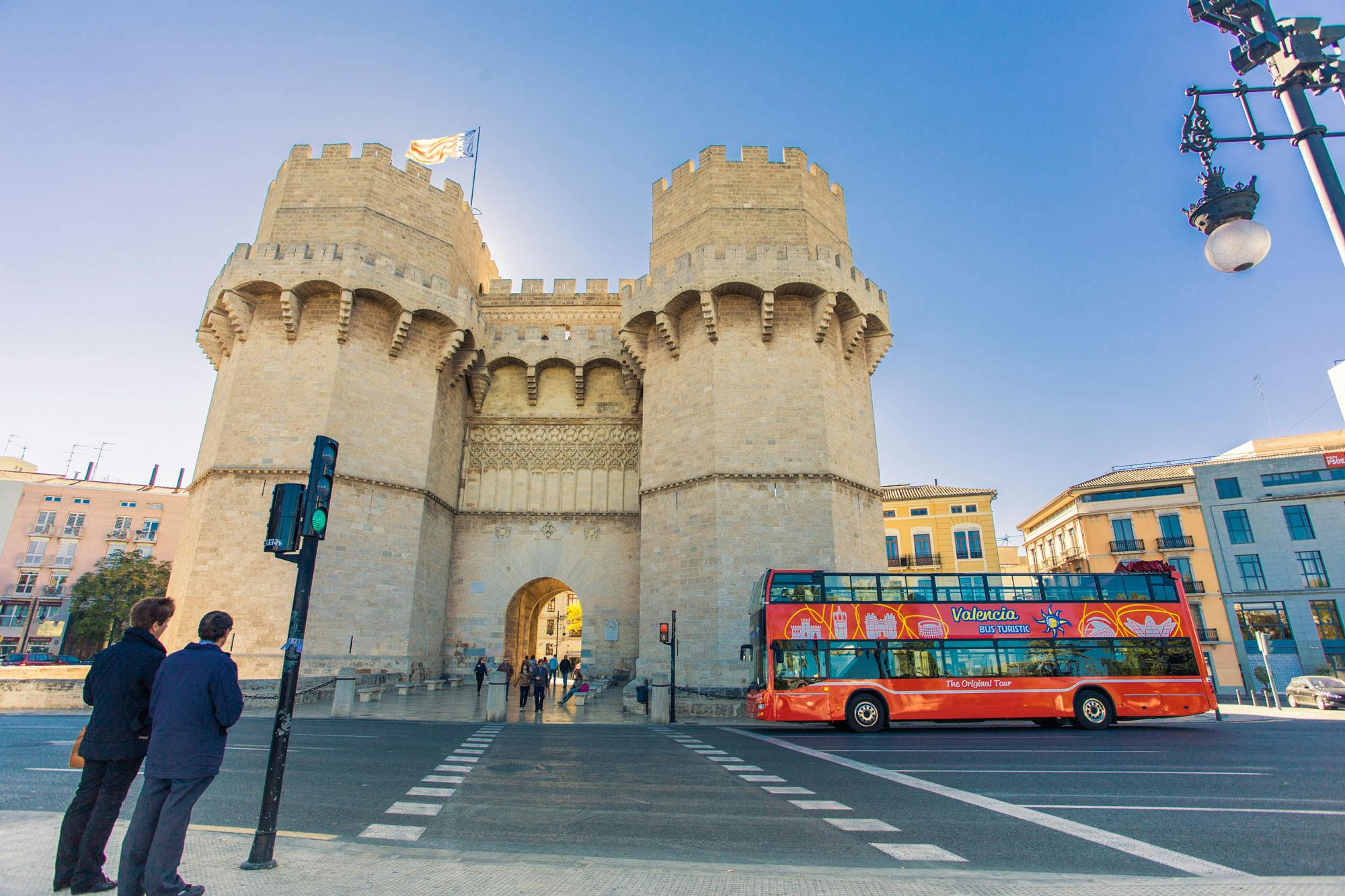 Stone castle gate with two towers, a red double-decker tour bus, and a person at a crosswalk under a clear blue sky.