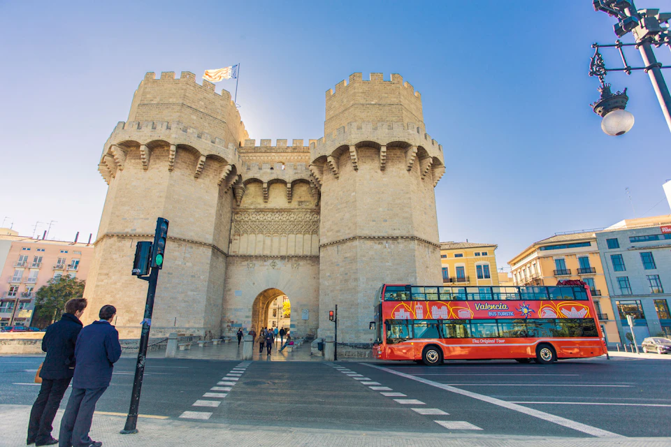 Stone castle gate with two towers, a red double-decker tour bus, and a person at a crosswalk under a clear blue sky.