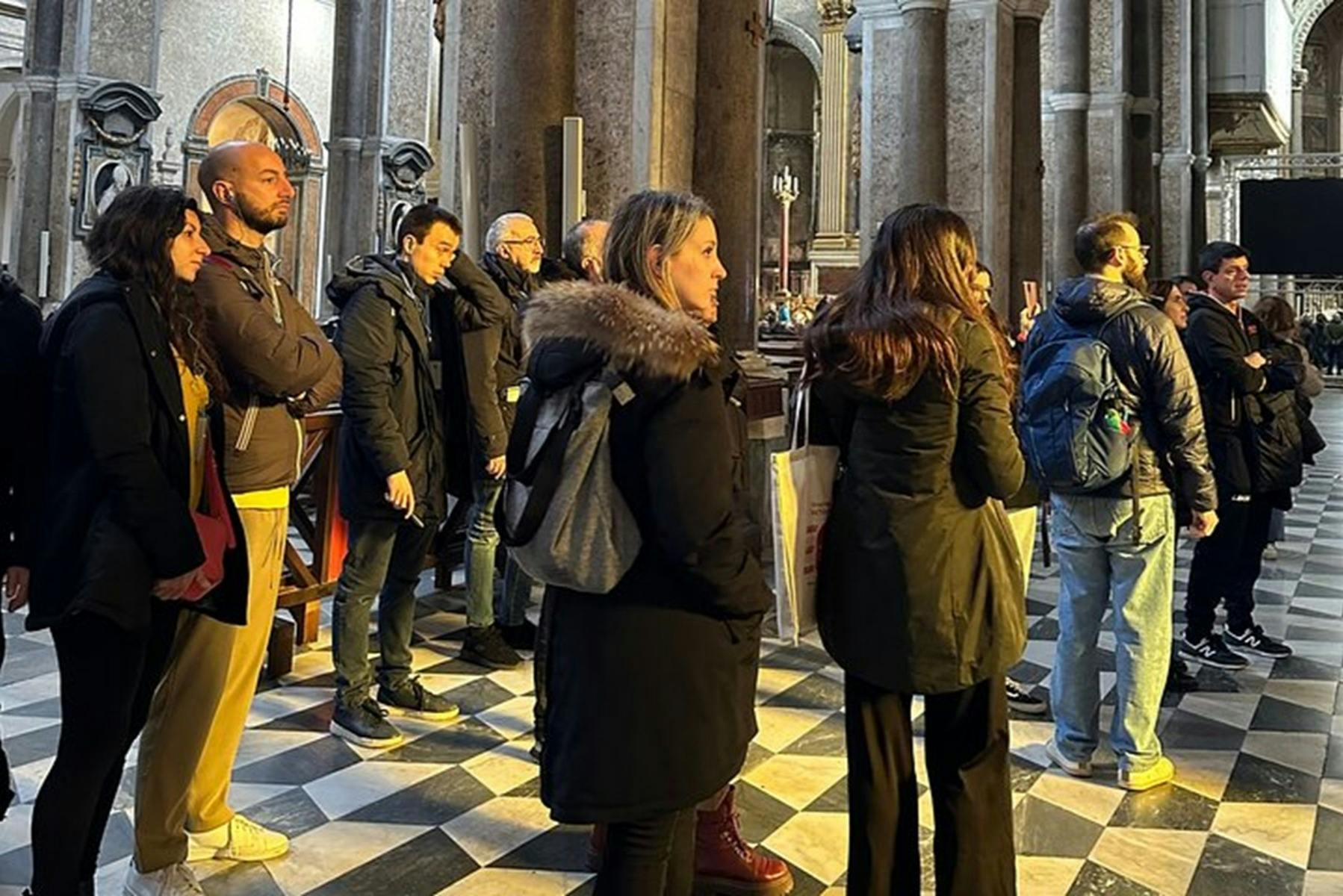Un grup de persones parades a l'interior sobre un terra amb patrons, envoltades de columnes de pedra i il·luminades per una llum càlida.
