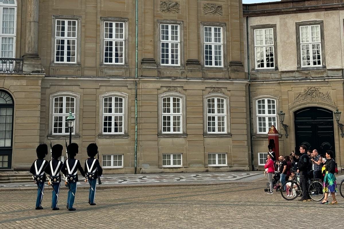 Guardie in uniforme e cappelli alti stanno davanti a un edificio storico, mentre alcune persone osservano da lontano.