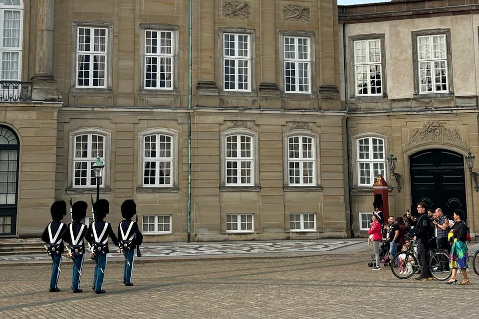 Guards in uniform and tall hats stand in front of a historic building, while a few people watch from a distance.