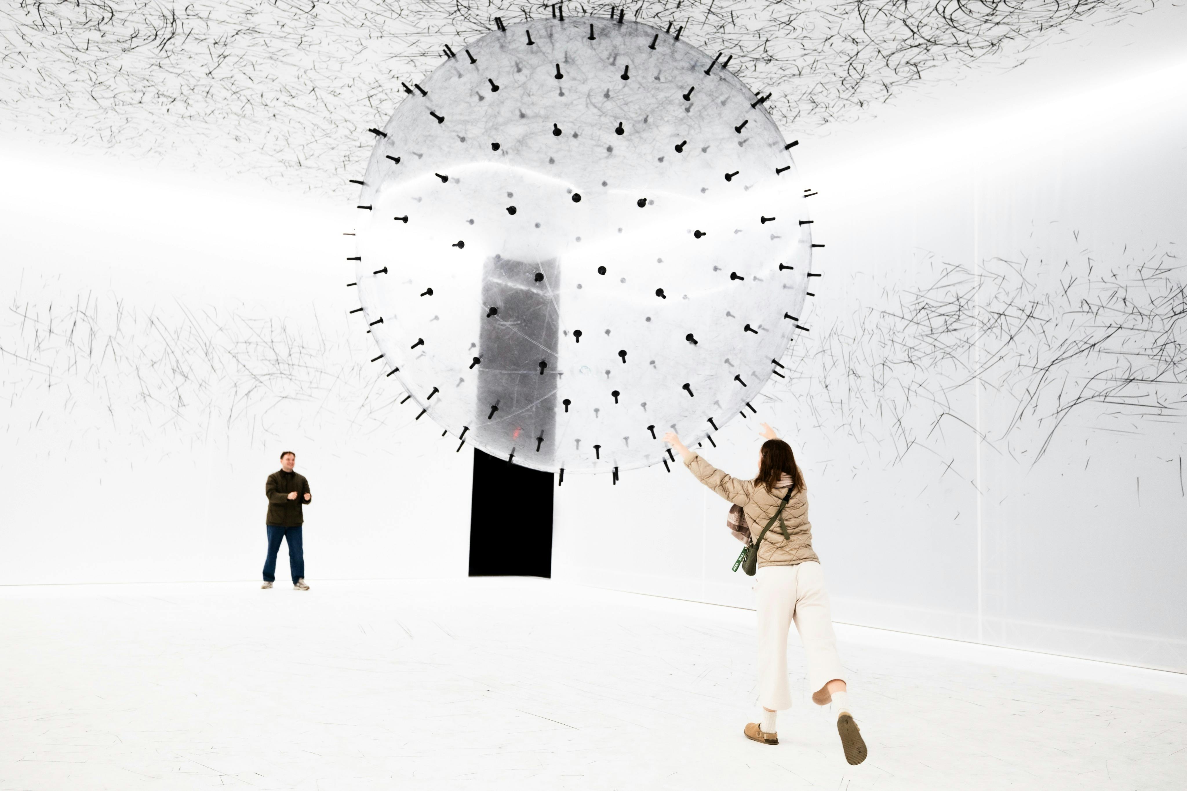 Человек взаимодействует с большой сферической художественной инсталляцией в белом пространстве галереи, в то время как другой человек наблюдает.