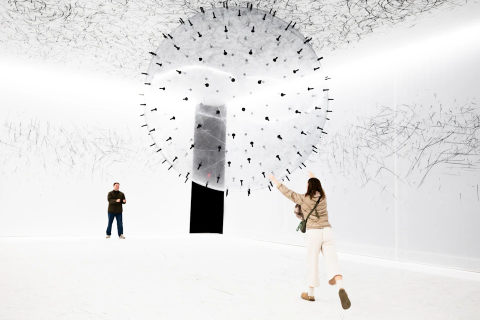 A person interacts with a large spherical art installation in a white gallery space, while another person observes.