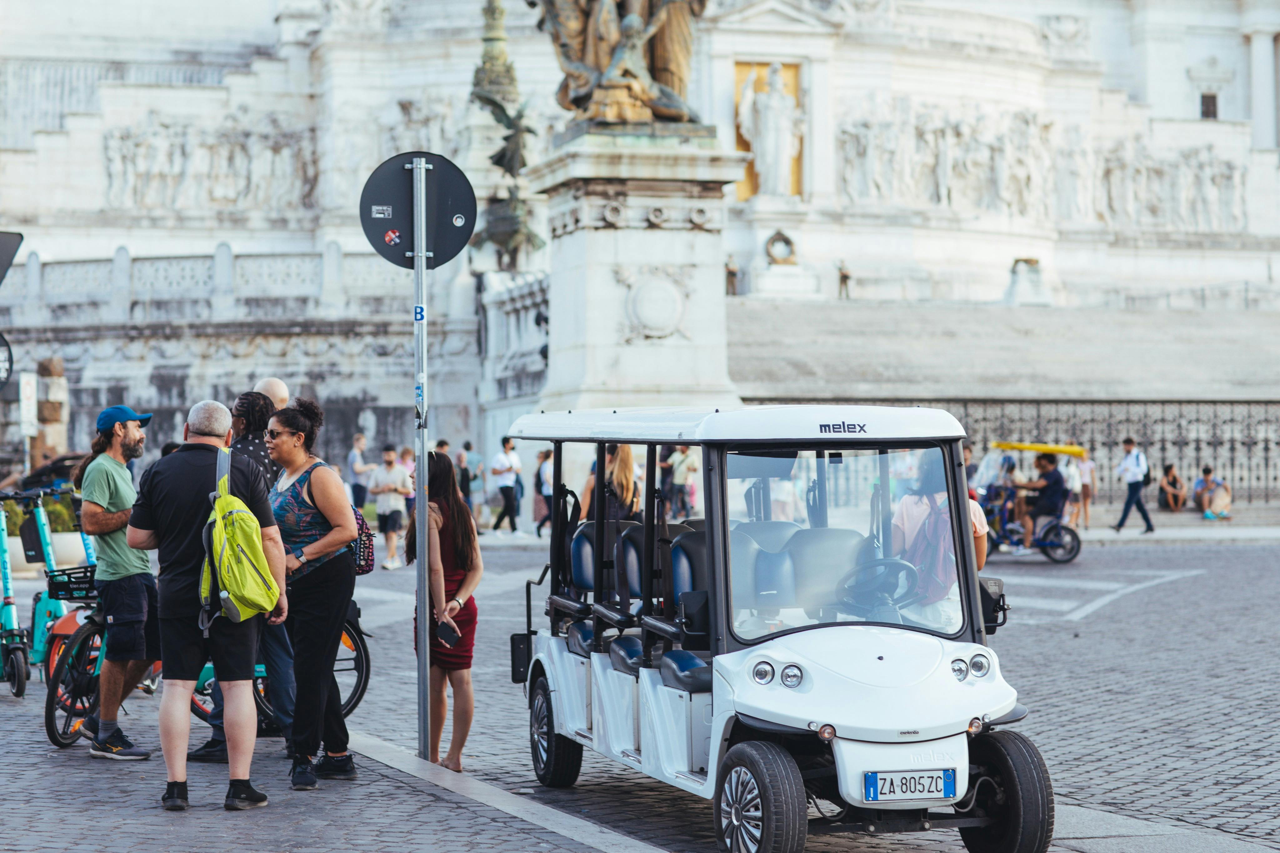 People standing and interacting near a white electric vehicle and bicycles in a historic urban square.