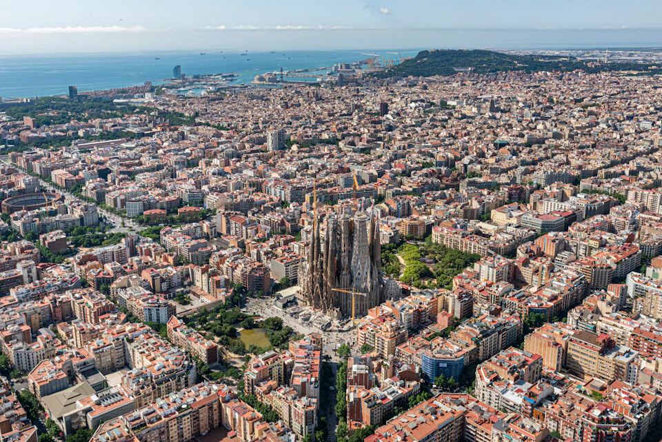 Aerial view of a large city with a prominent church at its center, surrounded by dense buildings and coastline in the distance.