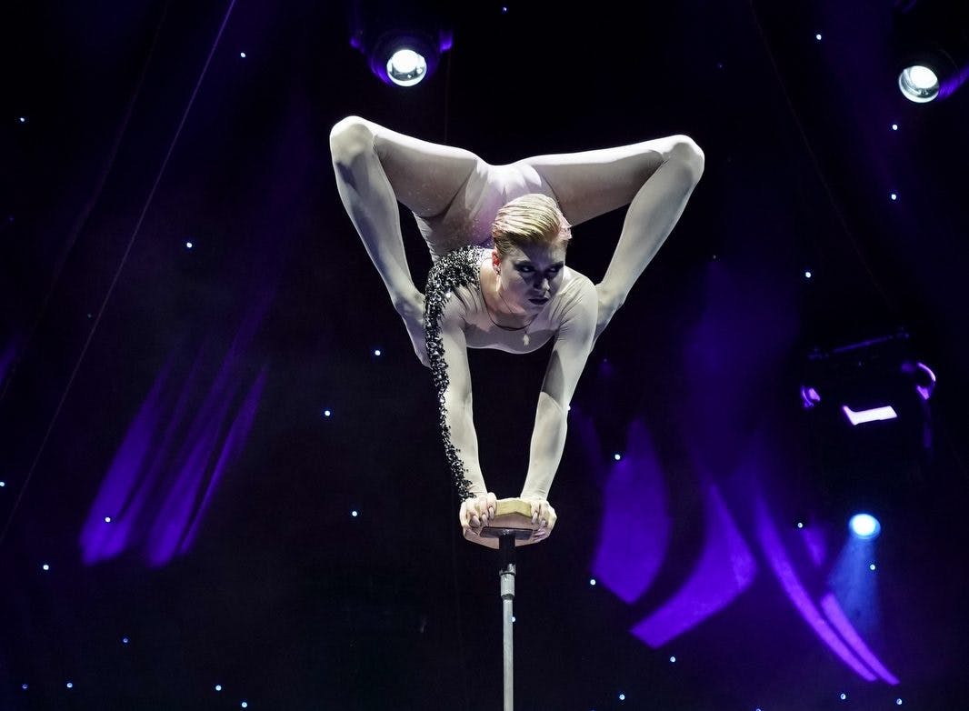 Acrobat i en vit dräkt utför ett handstående på en liten plattform med välvda ben, mot en scenbakgrund med lila belysning.