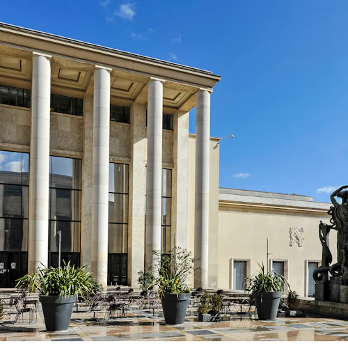 Exteriör av en neoklassisk byggnad med höga pelare, stora fönster, uteserveringar, krukväxter och en staty med ett spjut.