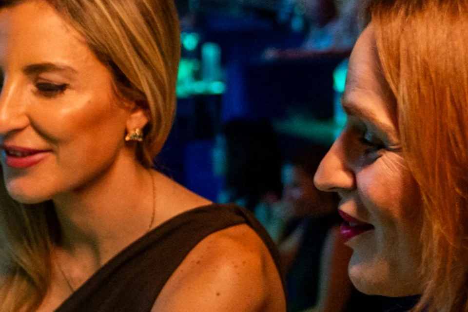Two women sitting at a table with plates of food and drinks, in a lively, dimly lit venue filled with people.