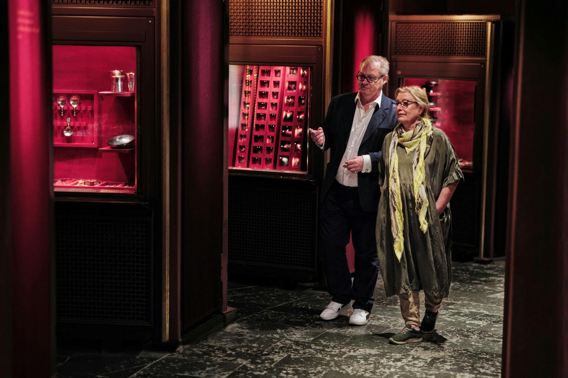 Två personer som går genom en svagt upplyst interiör med röda och guldfärgade displayer med olika föremål.