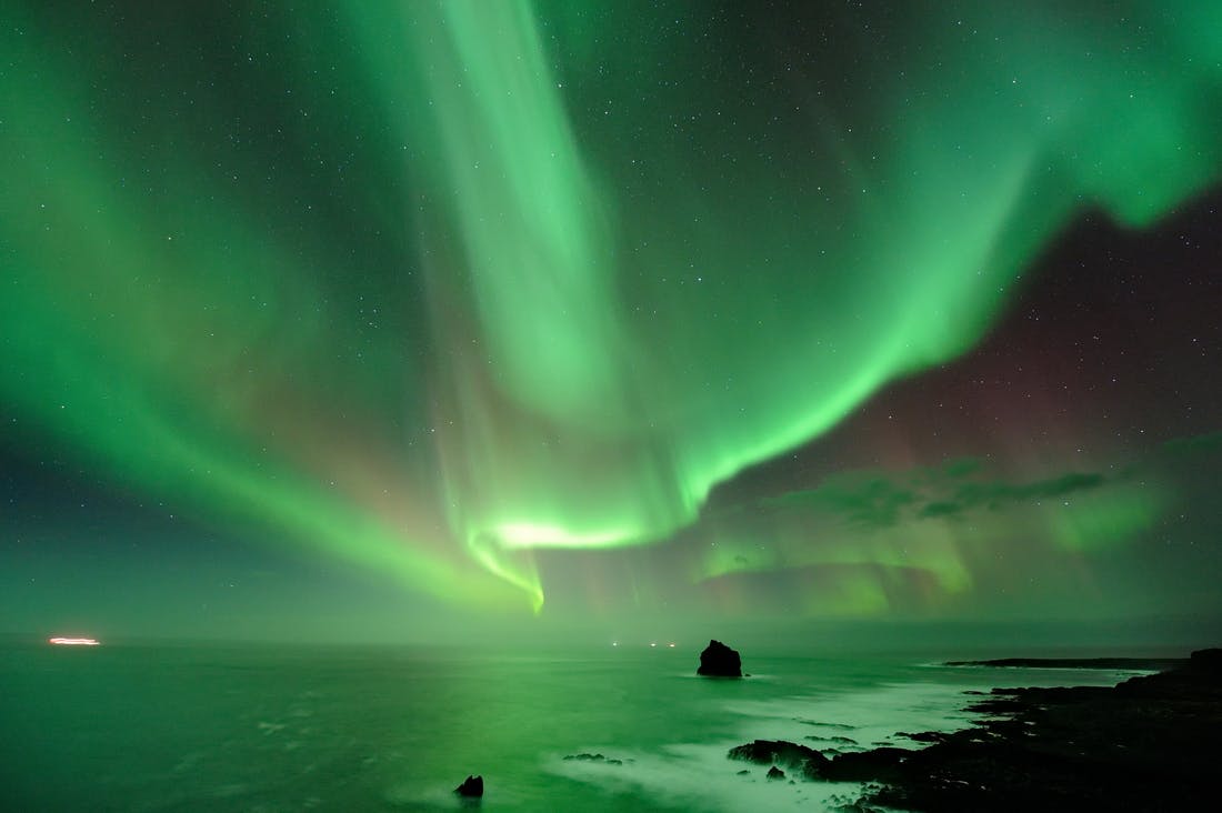 Aurora boreale verde brillante che illumina il cielo notturno su un paesaggio costiero con rocce e onde.