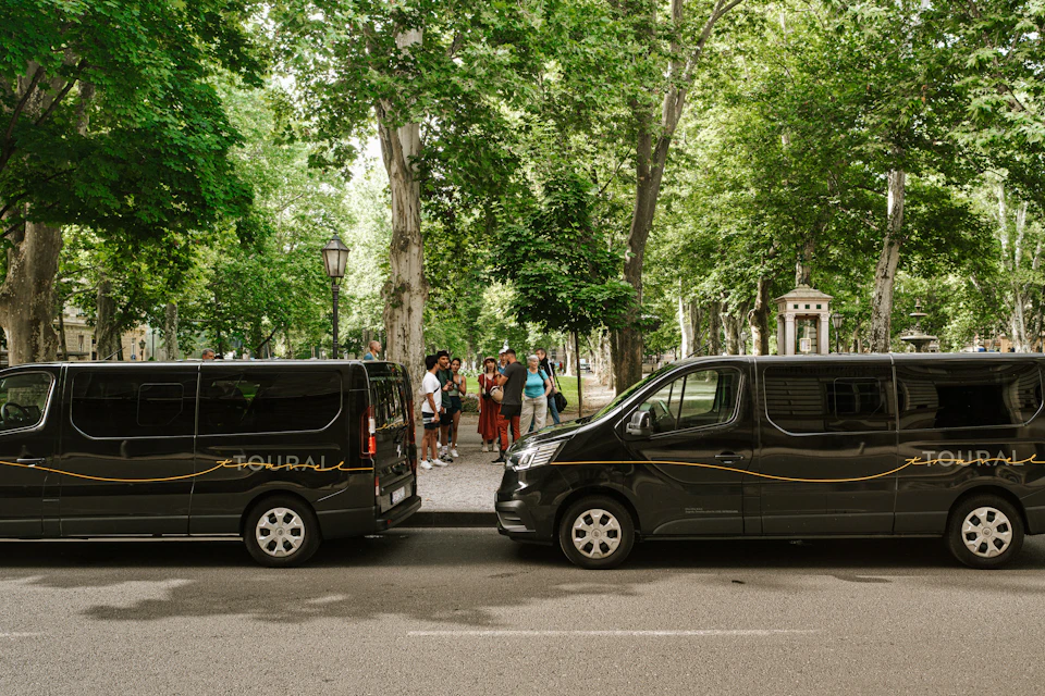 Two black vans parked on a street, with a group of people standing nearby in a park surrounded by trees.