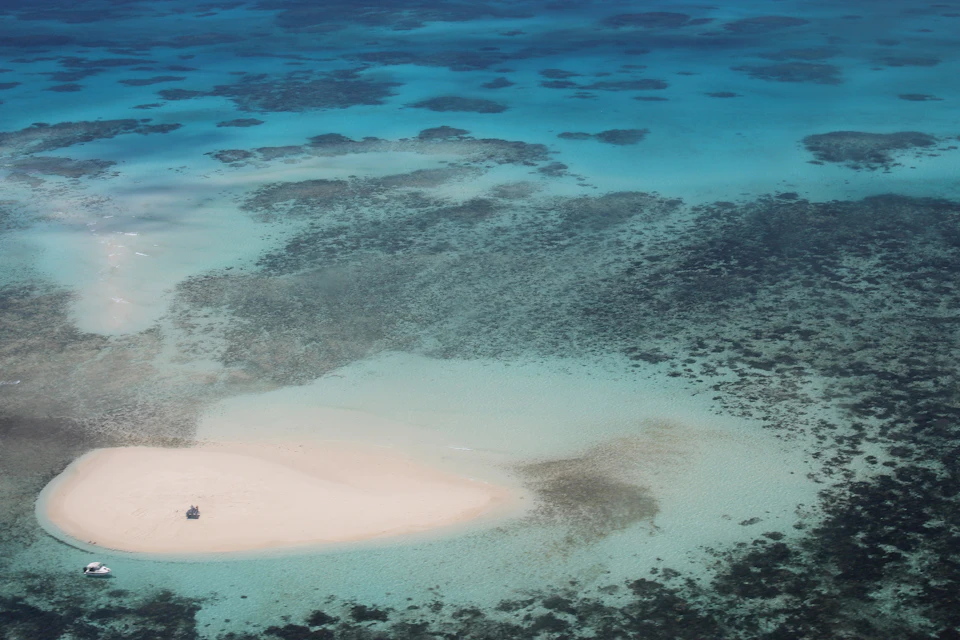 Aerial view of a remote sandy island surrounded by turquoise, shallow water with visible coral patches.