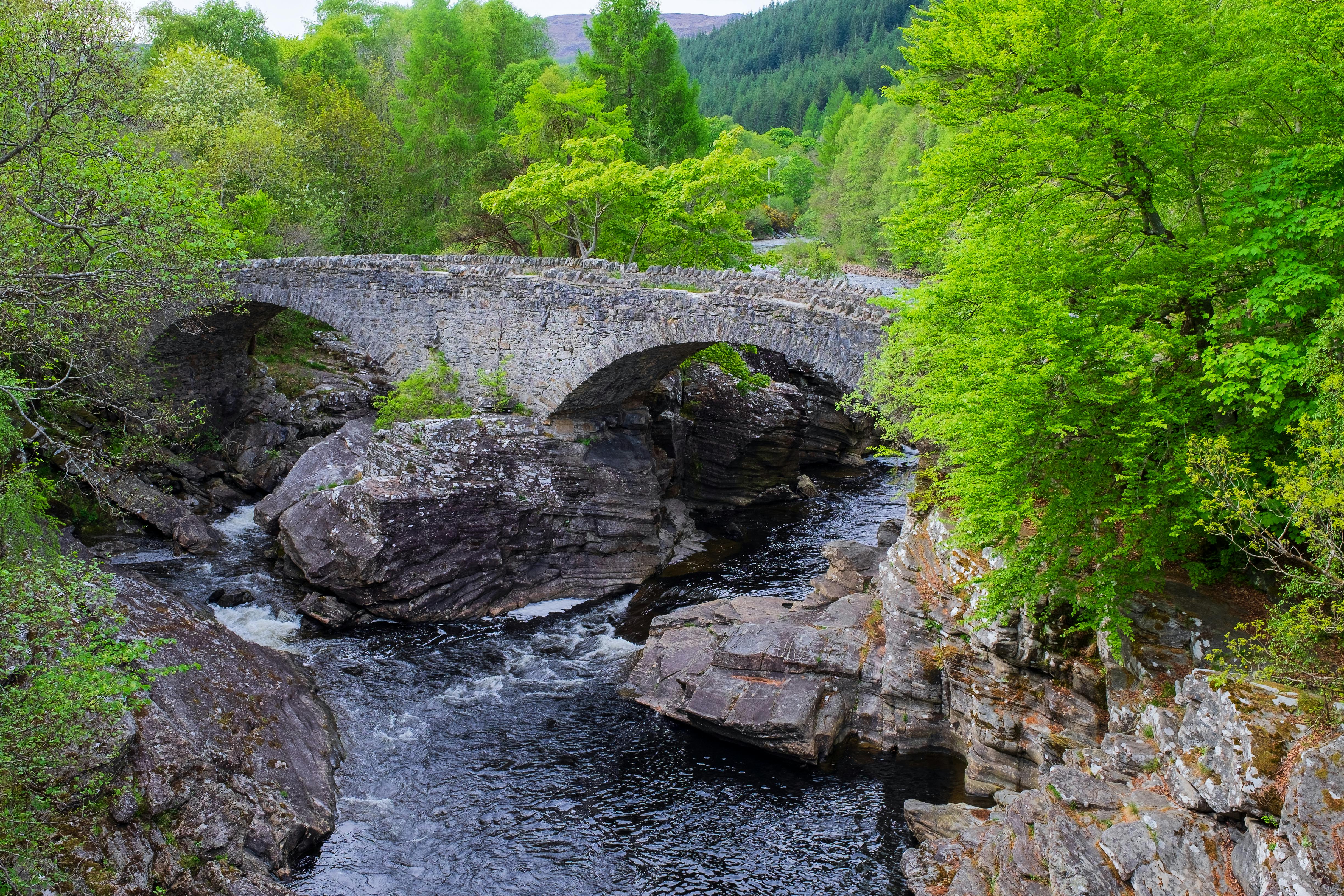 Μια πέτρινη γέφυρα καμαρώνει πάνω από ένα βραχώδες ρέμα που περιβάλλεται από καταπράσινα δέντρα και δάσος στο παρασκήνιο.