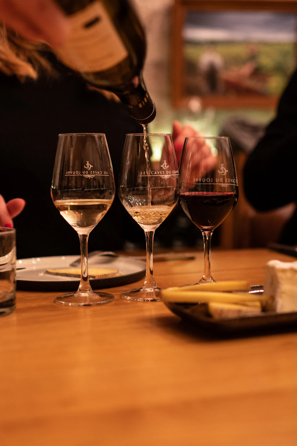 Three wine glasses on a wooden table, with wine being poured into one. Cheese and breadsticks are visible nearby.