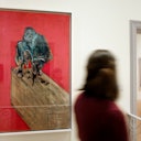 A person walks past a framed painting of a figure on a box against a red background in an art gallery.