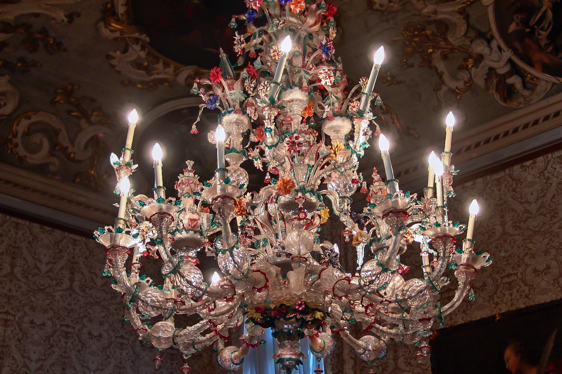 A large, ornate crystal chandelier with lit candles, adorned with colorful floral decorations, hangs from an elaborately decorated ceiling.