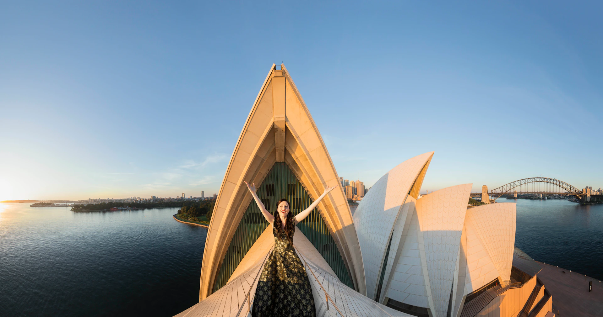 A woman in a patterned dress stands atop the Sydney Opera House, arms outstretched, with a view of the city and water in the background.