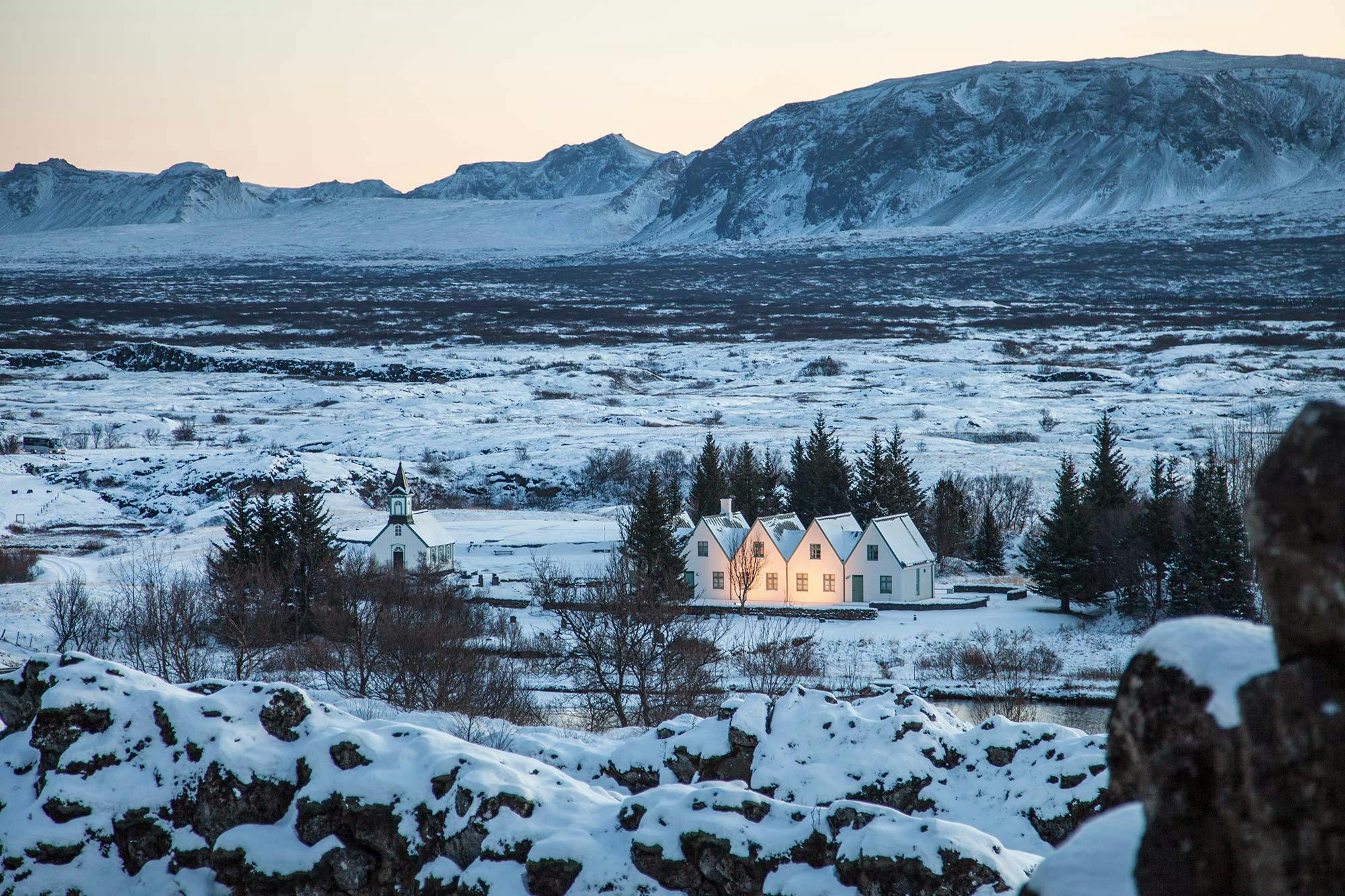 Snow-covered landscape with white houses, trees, and mountains in the background at dusk.