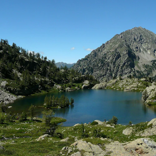 En klar fjällsjö omgiven av steniga, trädbevuxna sluttningar under en blå himmel, med ett stort berg i bakgrunden.