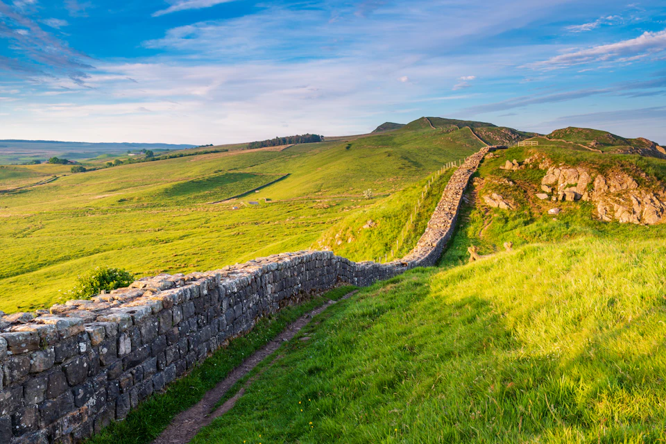 A stone wall winding through rolling green hills under a bright blue sky with scattered clouds.