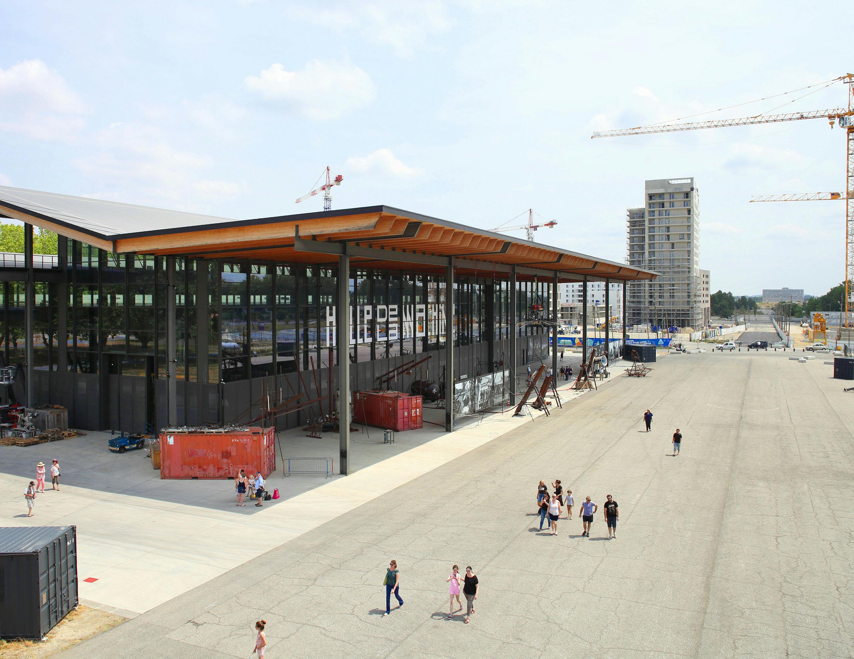 Люди гуляют рядом с современным зданием со стеклянными стенами и деревянной крышей, строительными кранами и высотными зданиями на заднем плане.