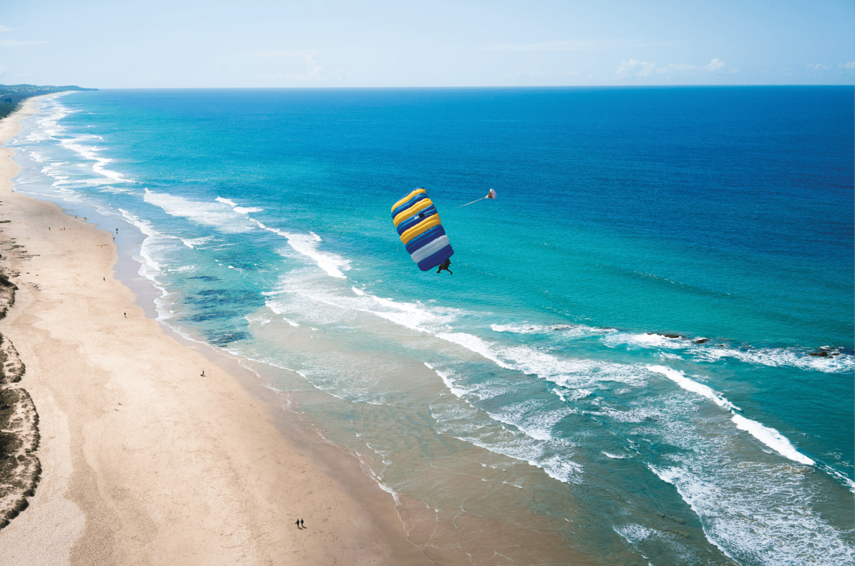 Person parasailing over blue ocean near sandy beach with small waves; a few people walk along the shore in the distance.