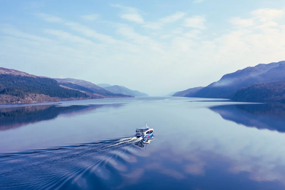 A boat sailing on a calm lake surrounded by mountains under a partly cloudy sky.