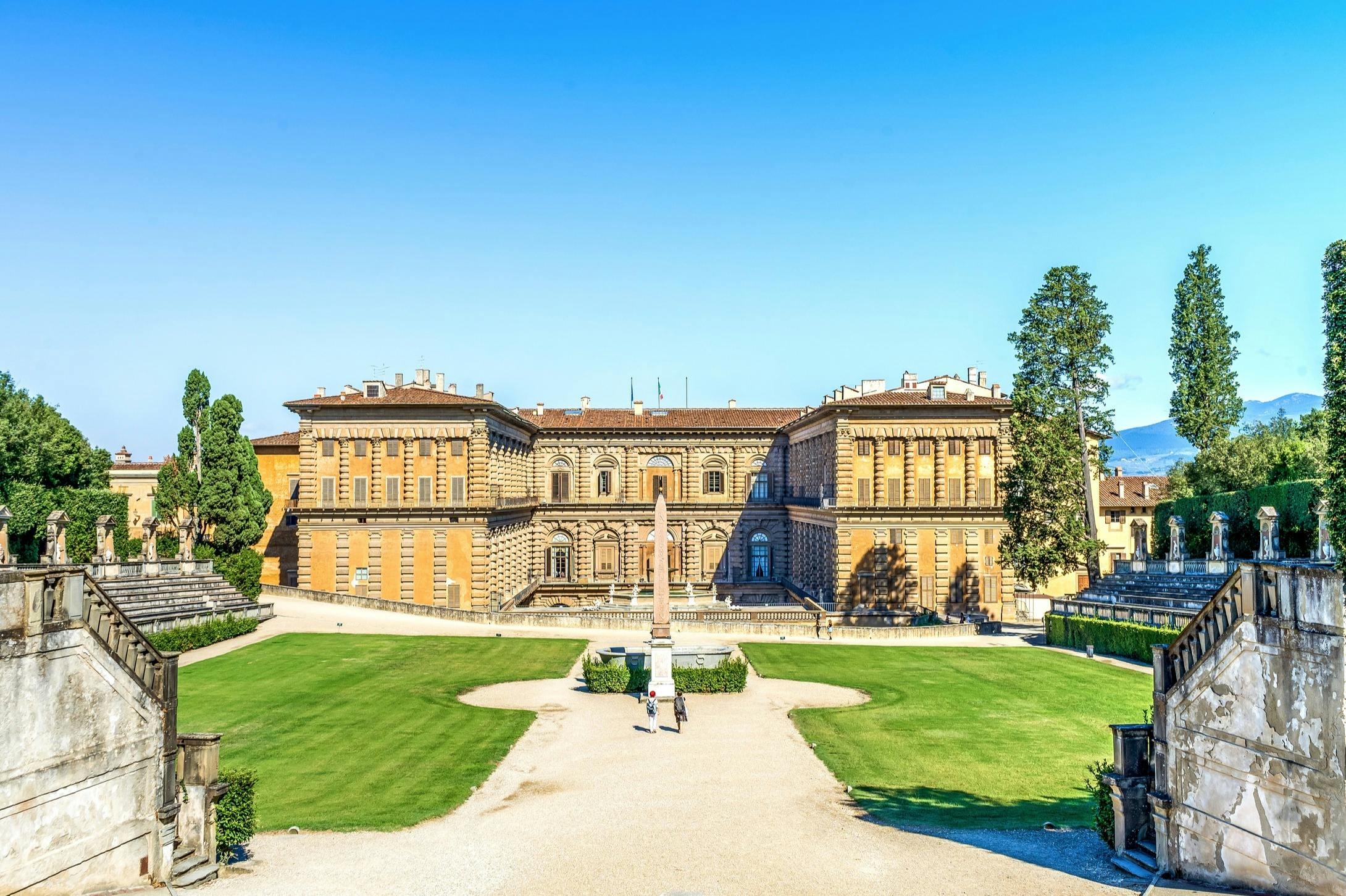 澄んだ青空の下、緑の芝生に囲まれた、対称的なファサードを持つ広大な歴史的建造物。 人々が道を歩いているのが見えます。