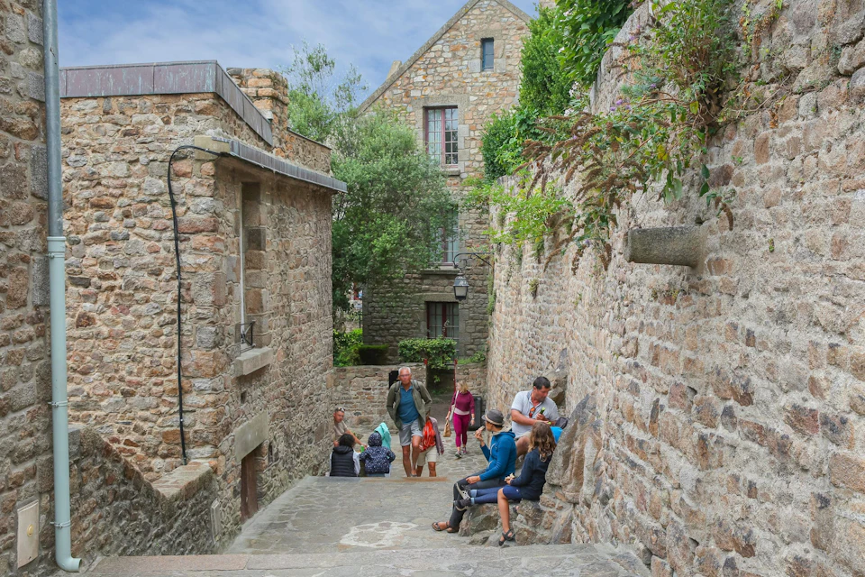 People sitting on stone steps and walking in a narrow alleyway within a historic stone village under a partly cloudy sky.