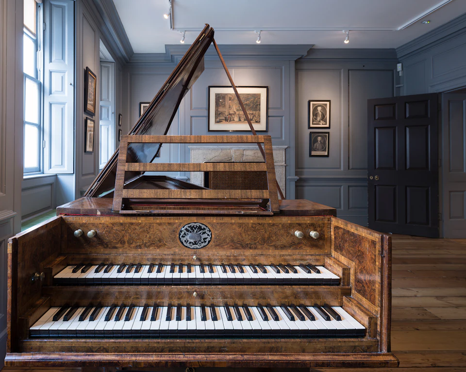 A harpsichord in a well-lit, elegant room with framed pictures on grey walls and wooden flooring.