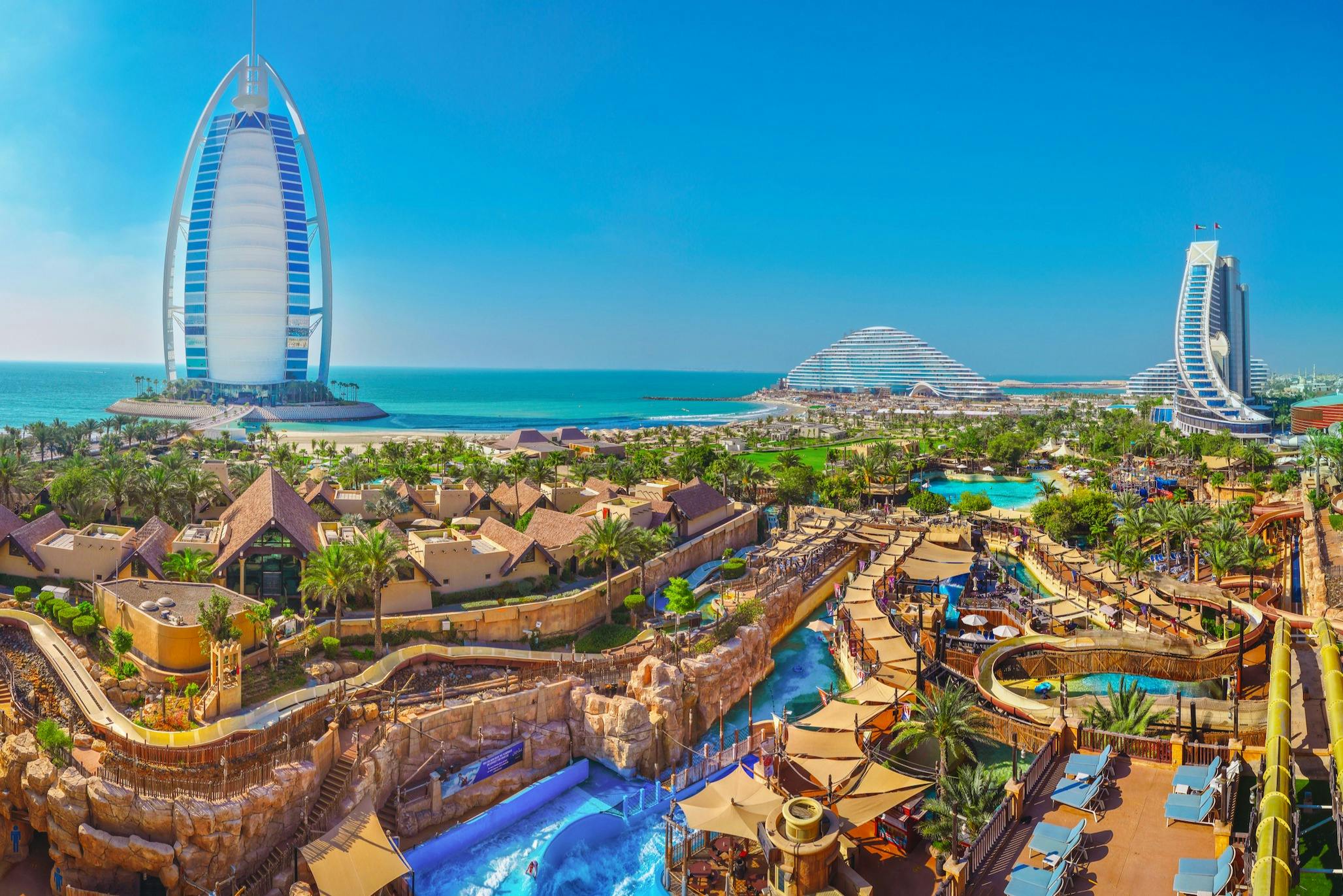 Un parc aquatique animé avec des toboggans, des piscines et des palmiers près de l'océan, avec des bâtiments modernes visibles à l'arrière-plan.