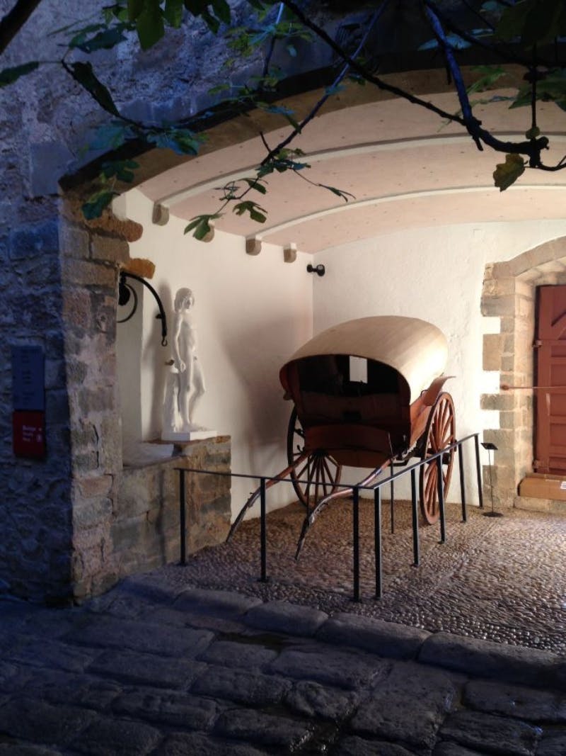 Под аркой выставлена старая деревянная тележка с брезентовым покрытием, с белой статуей слева и коричневой дверью справа.