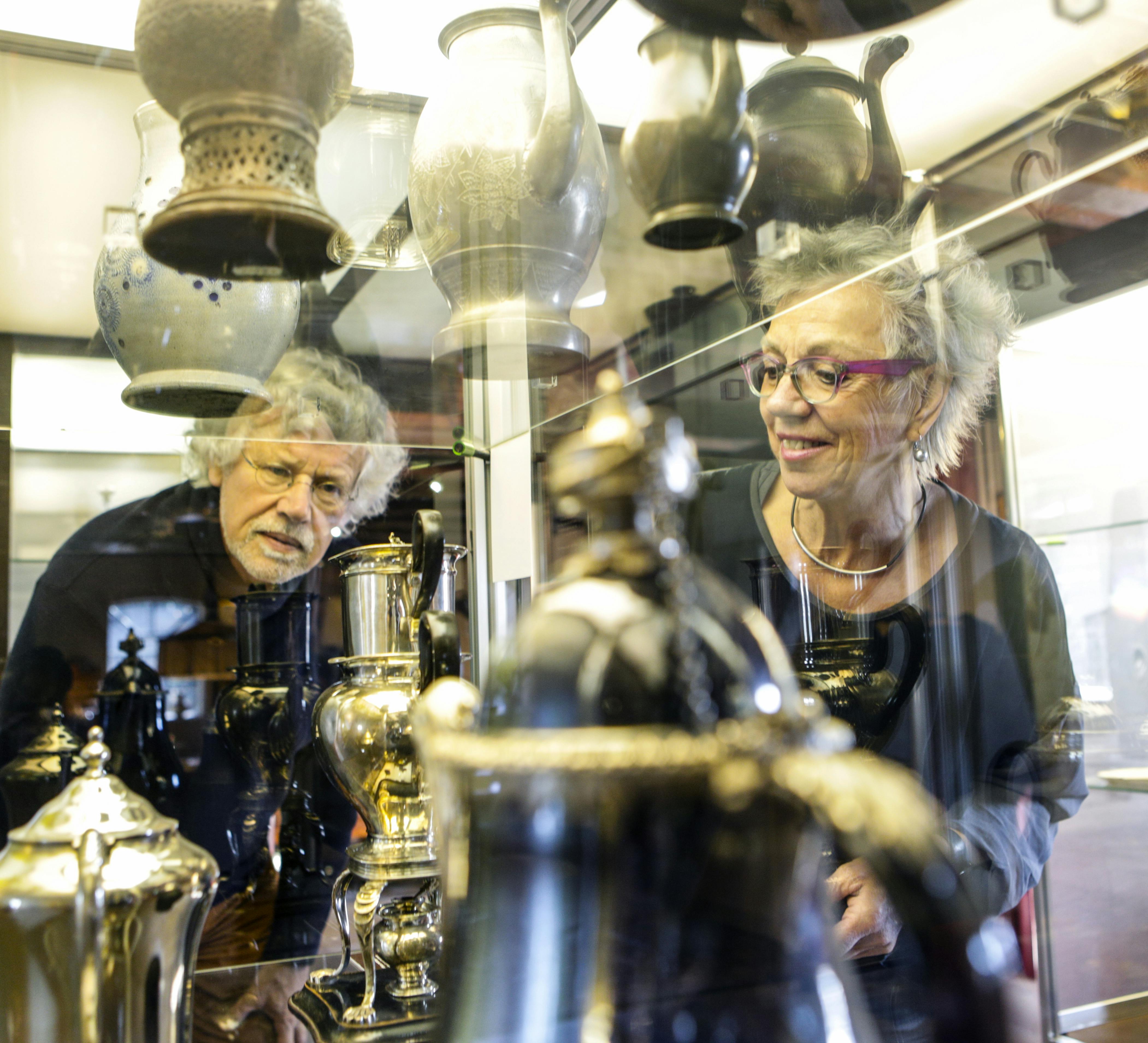 Два человека смотрят на витрину с антикварными вазами и горшками в стеклянном шкафу, отражающем их образы.