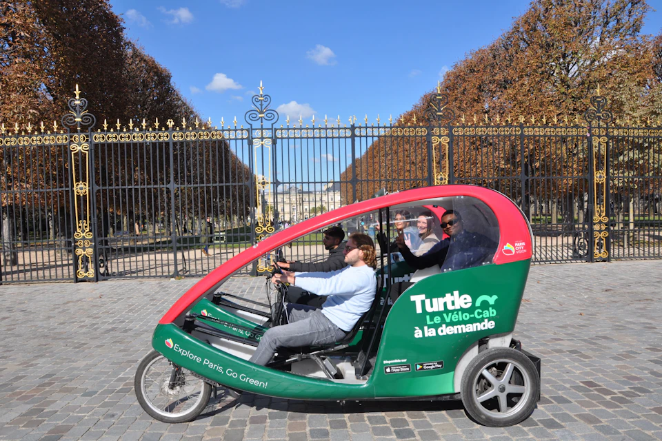 Four people sit in a green and red pedicab by "Turtle Le Vélo-Cab" in front of ornate black and gold gates on a sunny day.