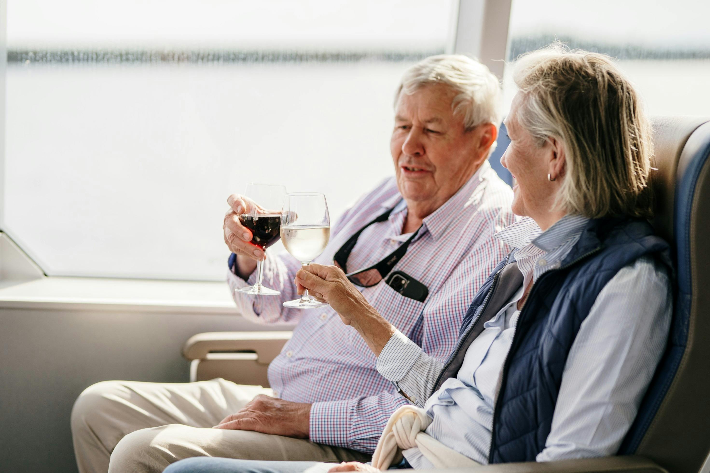 An older man and woman sitting by a window, holding glasses of wine and smiling at each other.