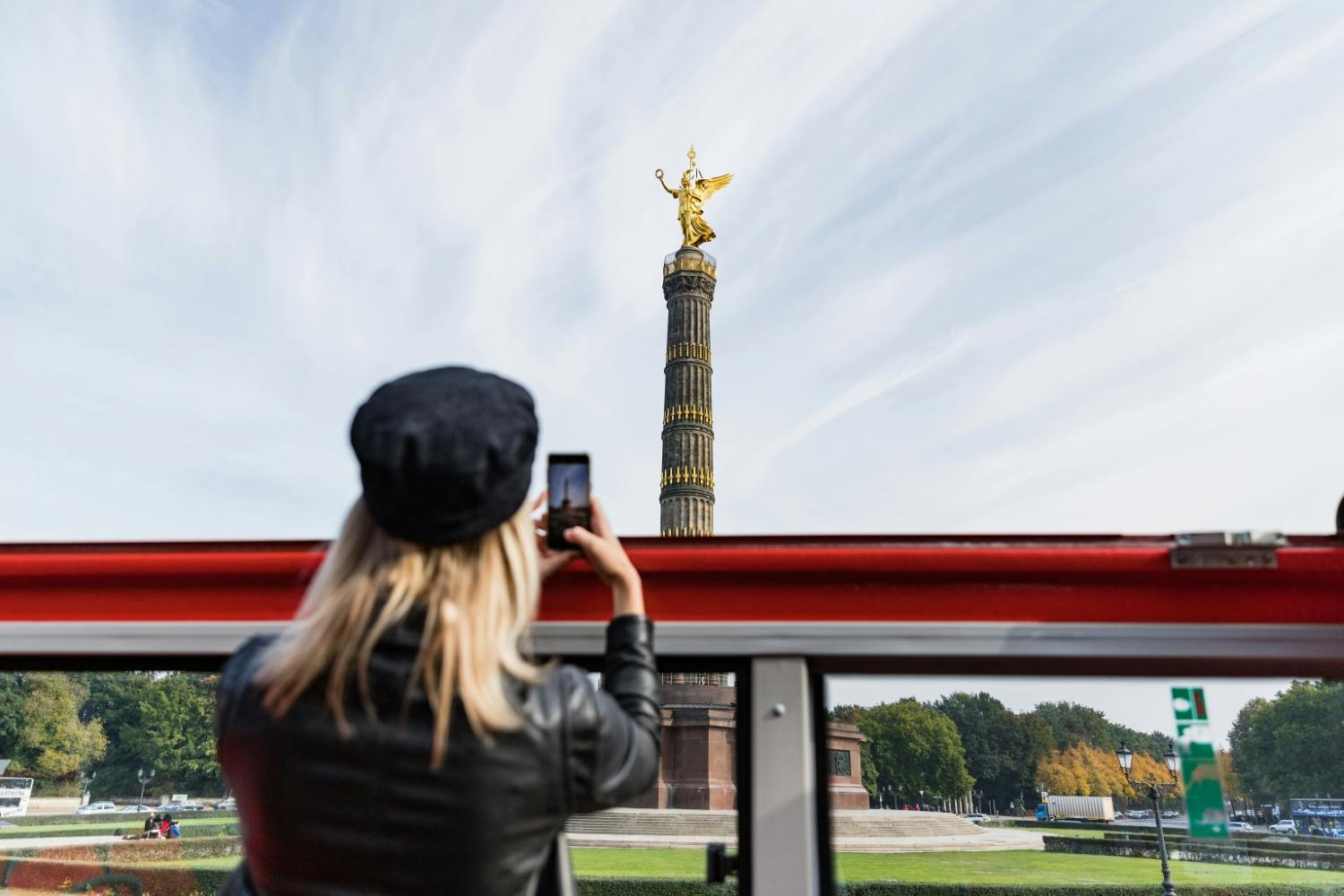 Una persona che indossa un berretto e una giacca nera fotografa un alto monumento con una statua d'oro in cima da un veicolo aperto.