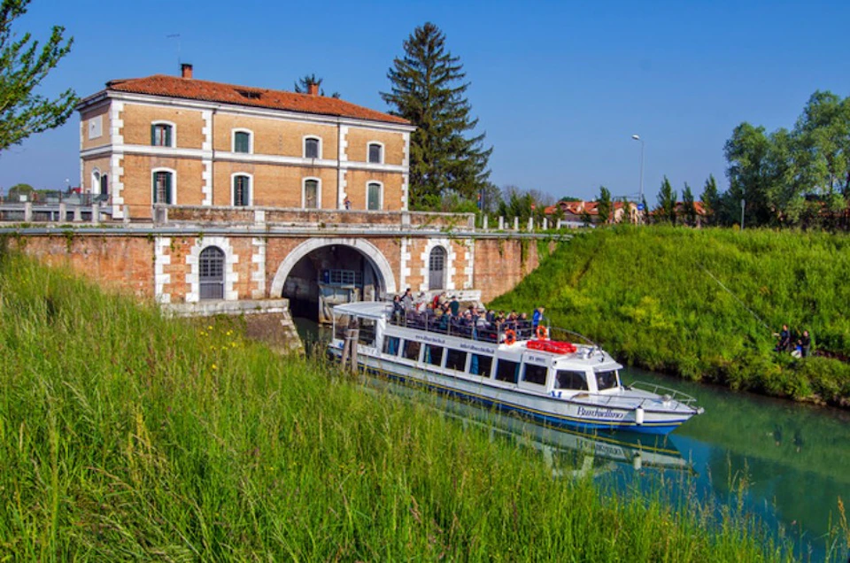 A boat with passengers enters a tunnel by a brick building with arched openings, surrounded by green grass and trees under a clear sky.