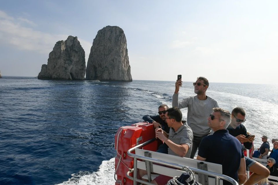 Four men on a boat near a rocky cliff. One is taking a selfie while others are seated, chatting, and enjoying the view.