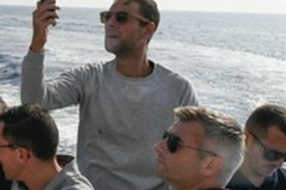 Four men on a boat near a rocky cliff. One is taking a selfie while others are seated, chatting, and enjoying the view.