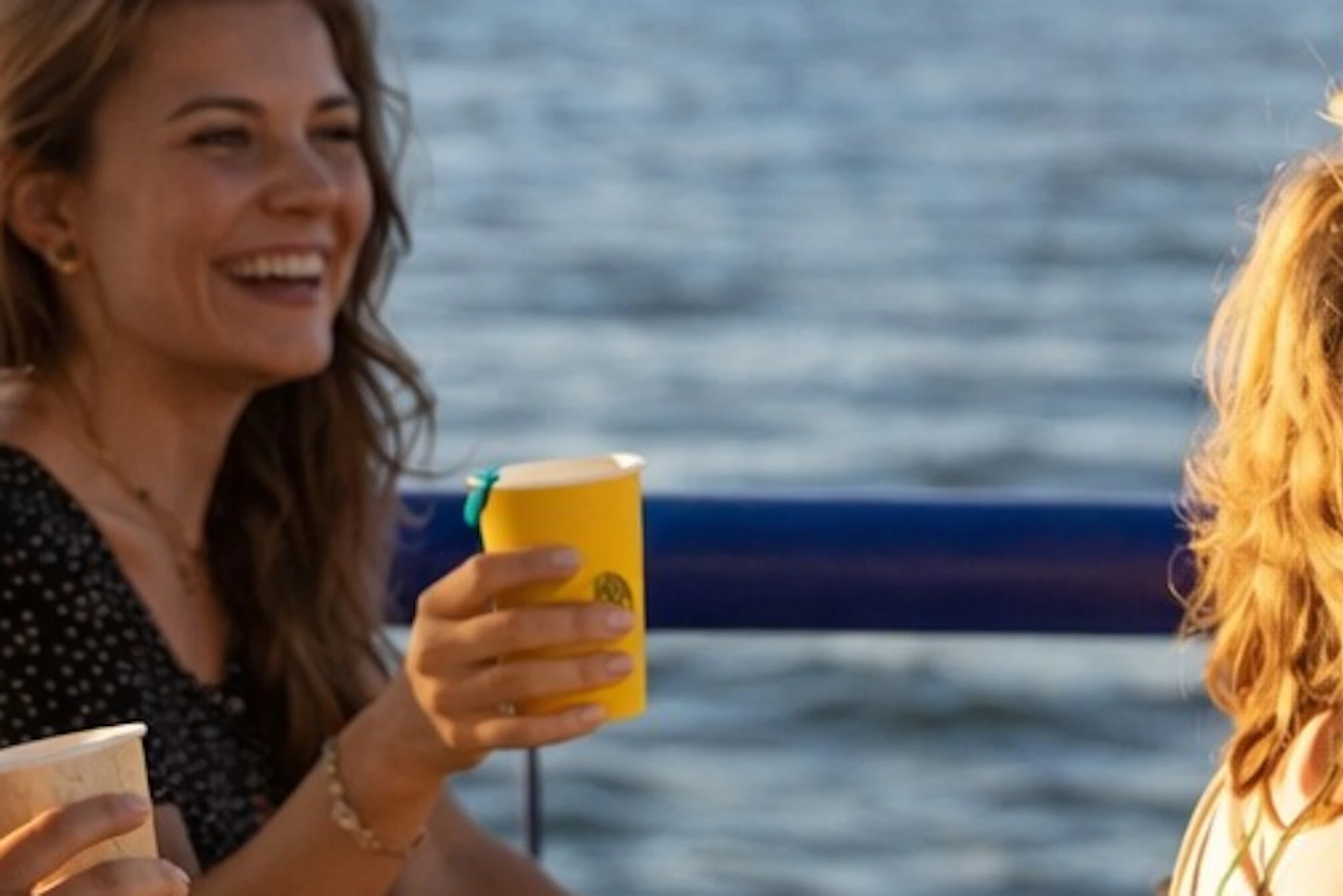 Three people sitting on a boat, holding drinks and enjoying a sunny day with water and a cityscape in the background.