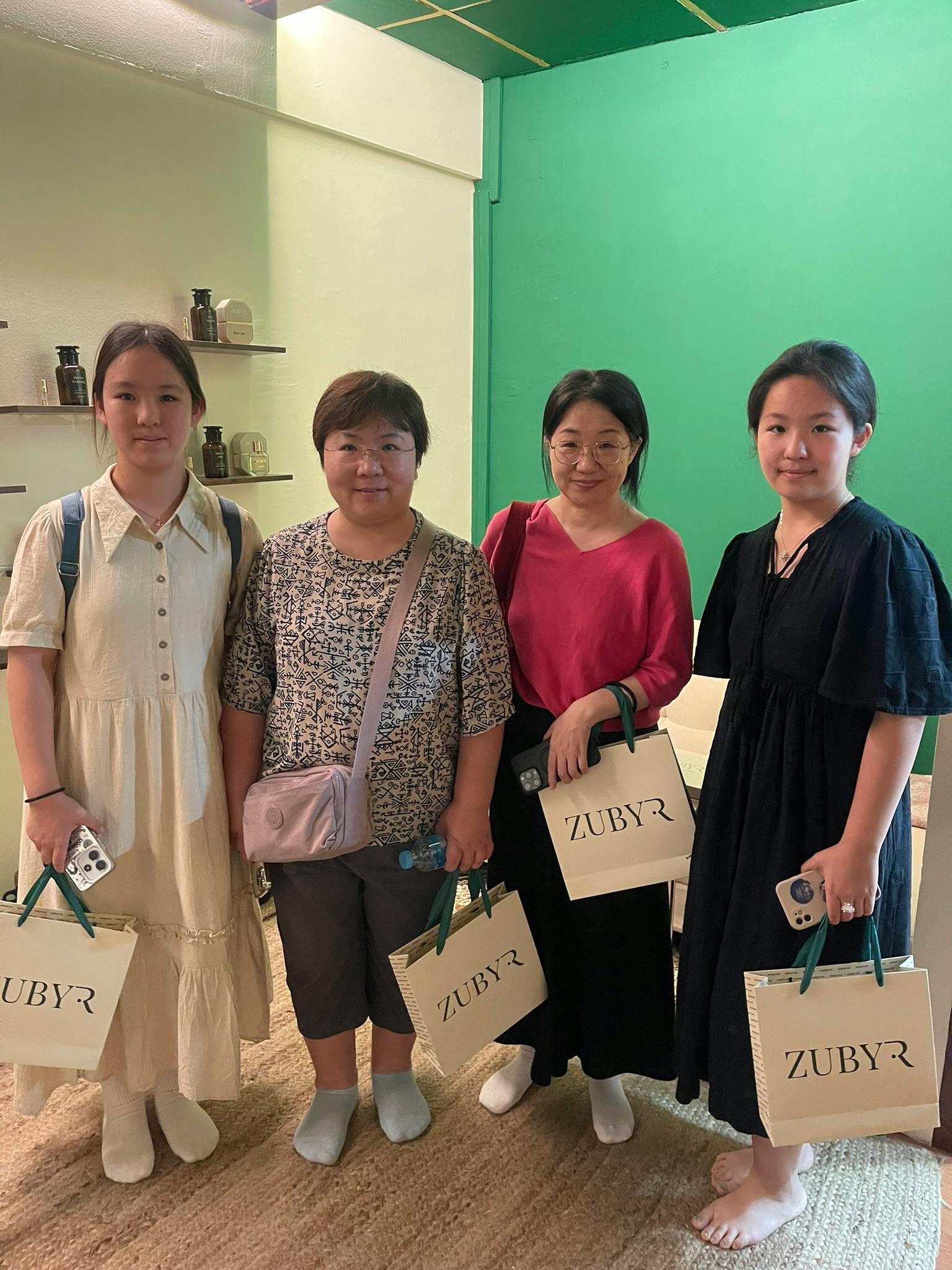 Four women standing indoors near a green wall, holding shopping bags and posing for a photo.